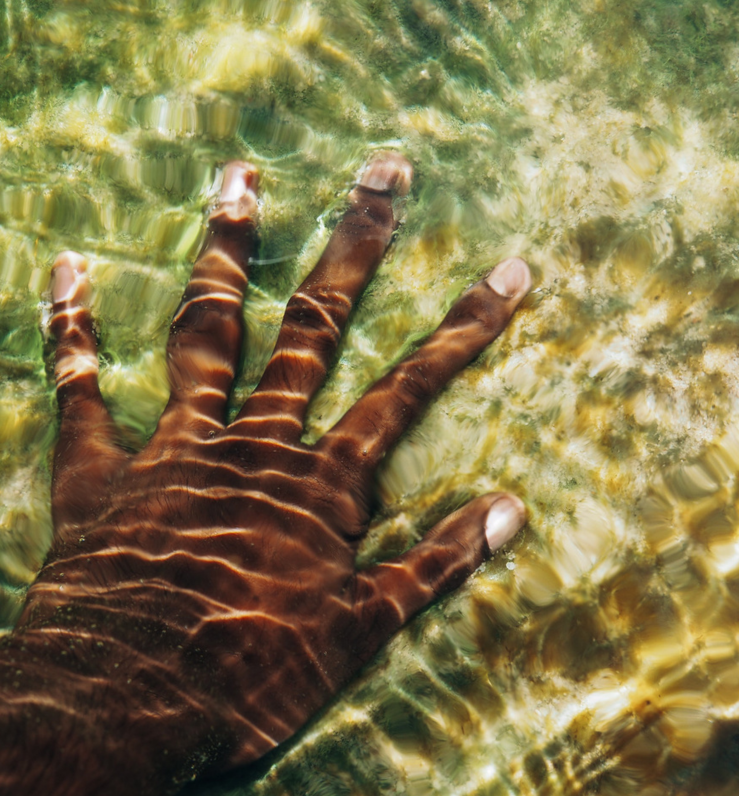 A close-up of a hand submerged in clear, shallow water, with sunlight creating rippling patterns across the skin and the sandy, green-tinted surface below.